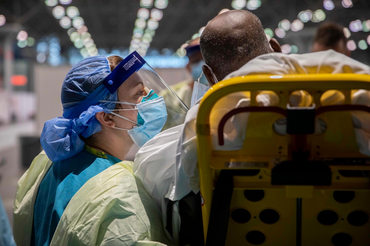 A medical professional wearing protective gear leans over to speak to a patient laying in a bed.