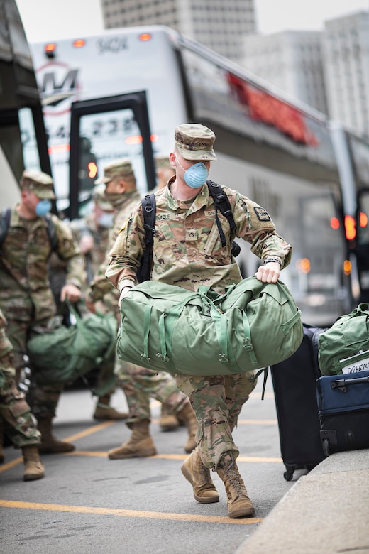 Soldiers in uniform and face masks carry duffle bags off a bus.