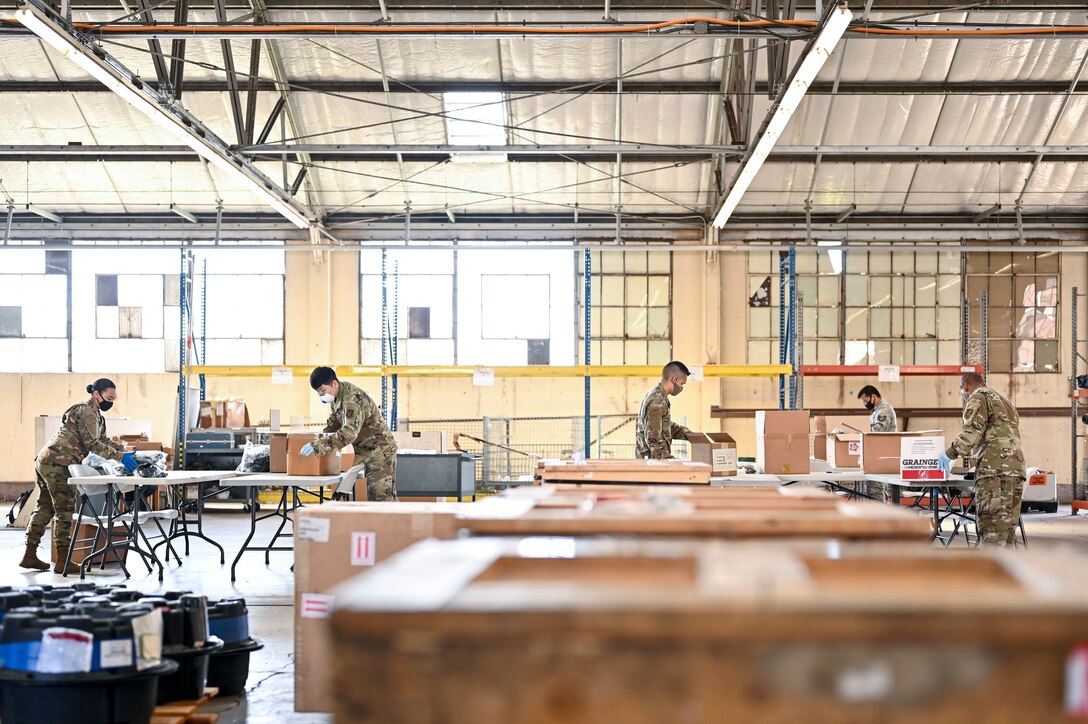 Airmen stand and work at tables in a large room.