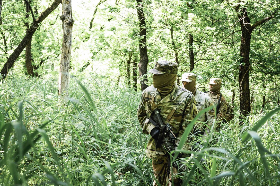 Soldiers wearing face masks walk through a wooded area.