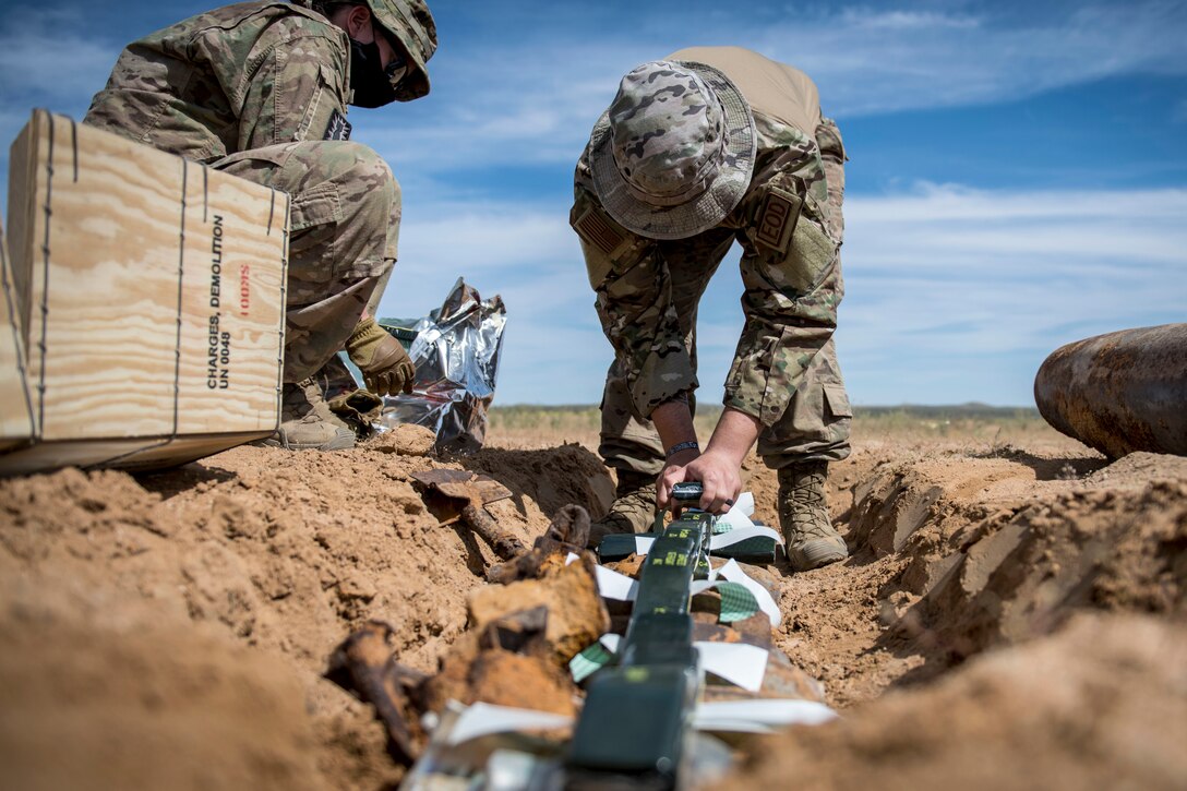 An airman arranges ordnance in a dirt pit as another observes.