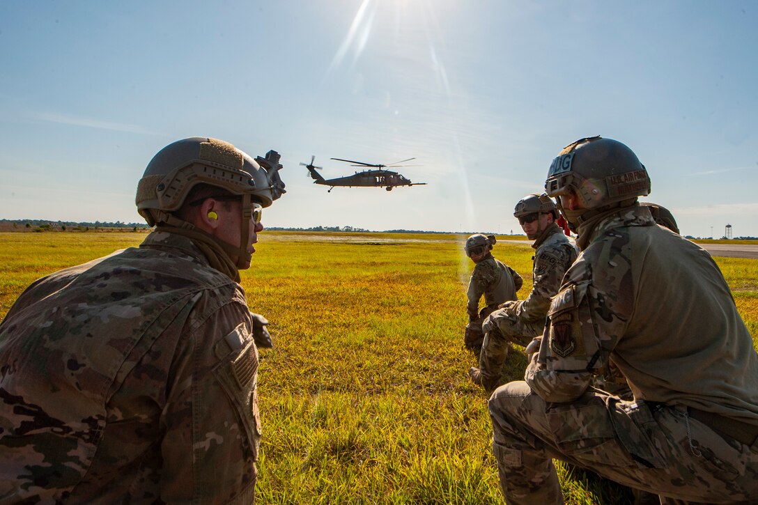 A photo of airmen talking while a helicopter flies in the background.