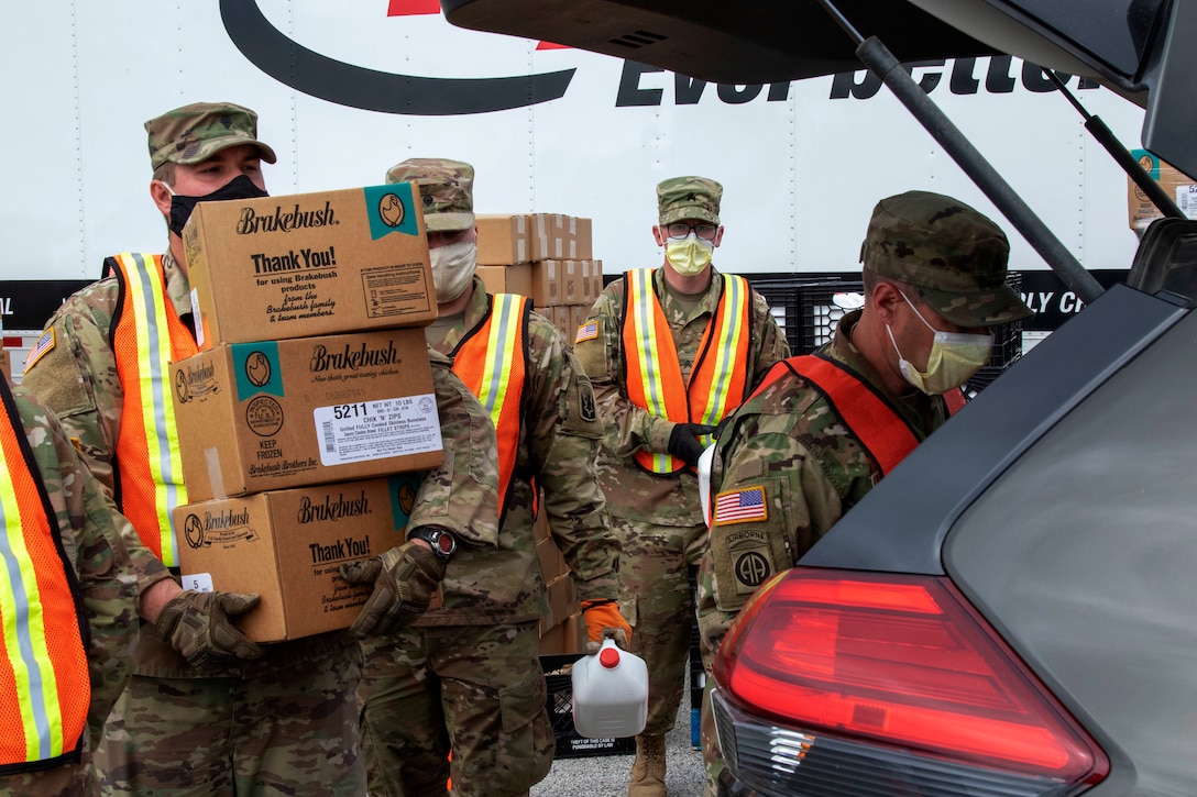 Four soldiers wearing face masks and gloves carry boxes of packaged food to the vehicles.