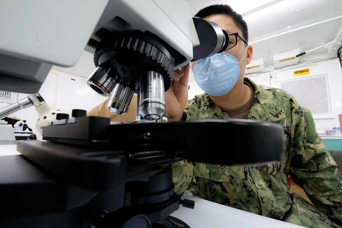 A sailor in protective gear looks through a microscope.
