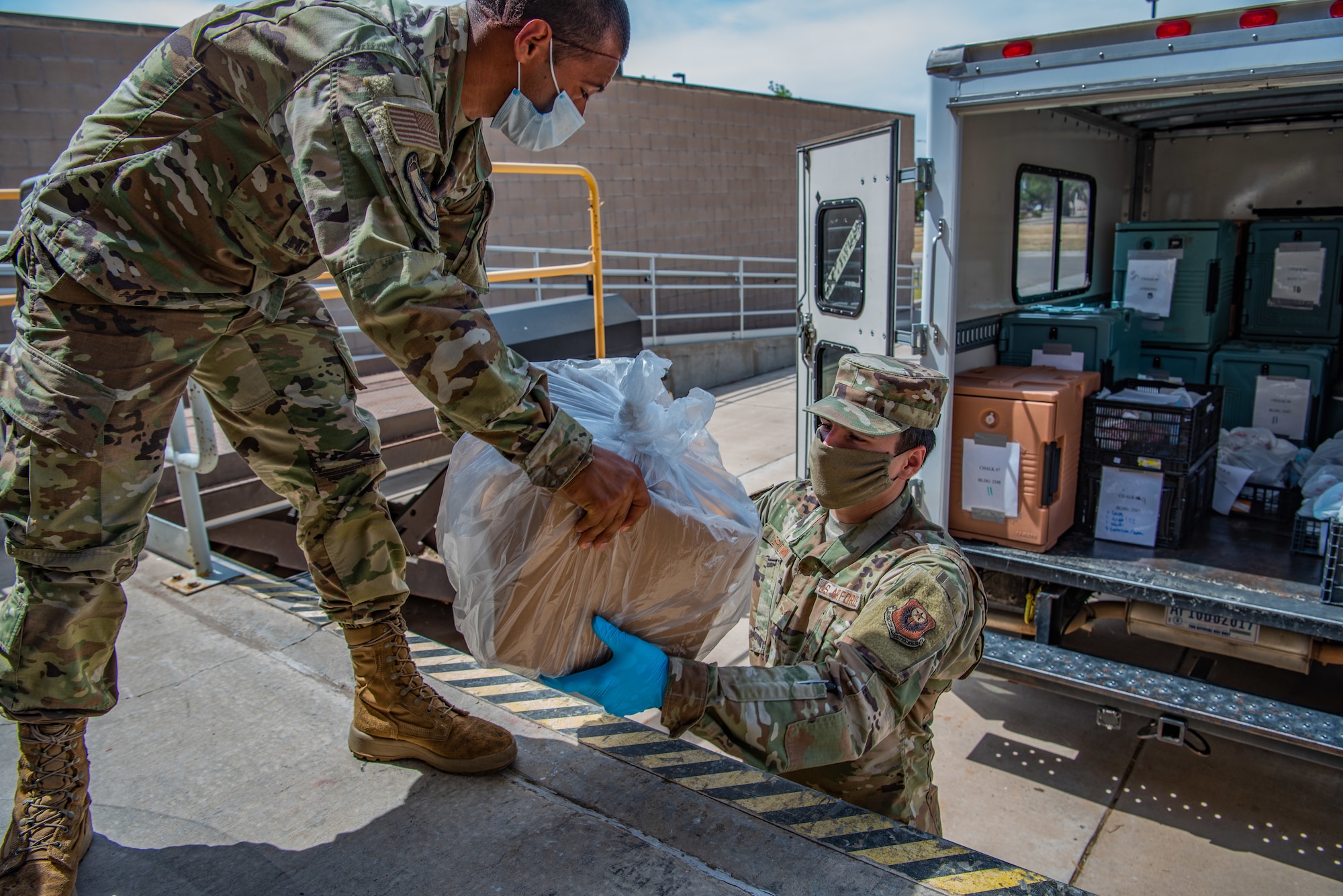 Airman 1st Class Gabrield Alvarado, left, a food services specialist assigned to the 27th Special Operations Force Support Squadron, hands a box of food to Airman 1st Class Brandon Brown, a ground transportation apprentice assigned to the 27th Special Operations Logistics Readiness Squadron, for delivery to quarantined Airmen at the Pecos Trail Dining Facility on Cannon Air Force Base, N.M., May 15, 2020. Each hot meal delivered to Airmen quarantined to the base contains a starch, a vegetable or fruit, a protein and a bread, as well as drinks and snacks. (U.S. Air Force photo by Senior Airman Maxwell Daigle)