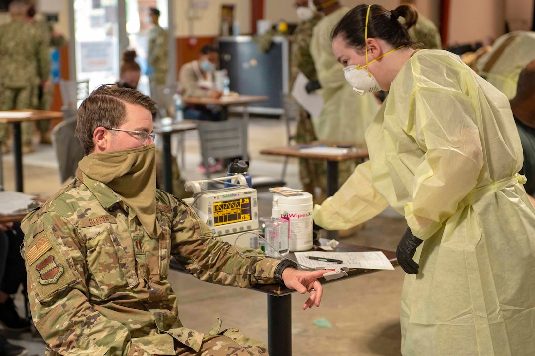 A sailor wearing personal protective equipment checks vitals on a seated service member.