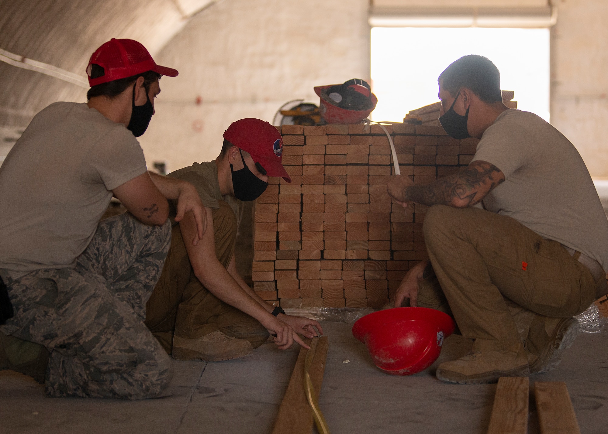 Two RED HORSE Airmen watch as another RED HORSE Airman uses a measuring tape to measure a piece of wood.