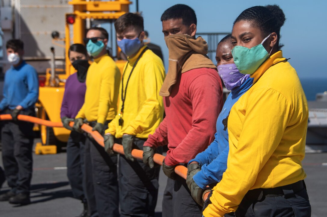 Sailors stand in a row holding a long firefighting hose.