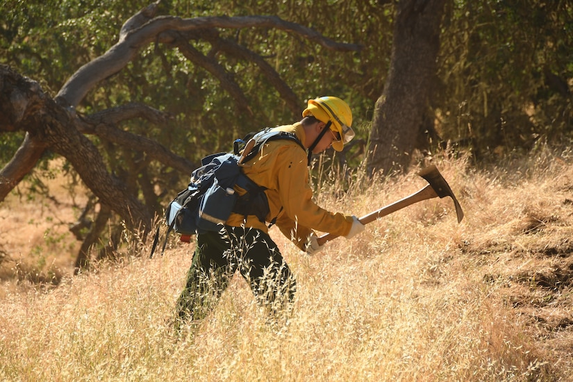 A soldier wearing fire-protection equipment uses a hand tool in a field of tall, dry grass.