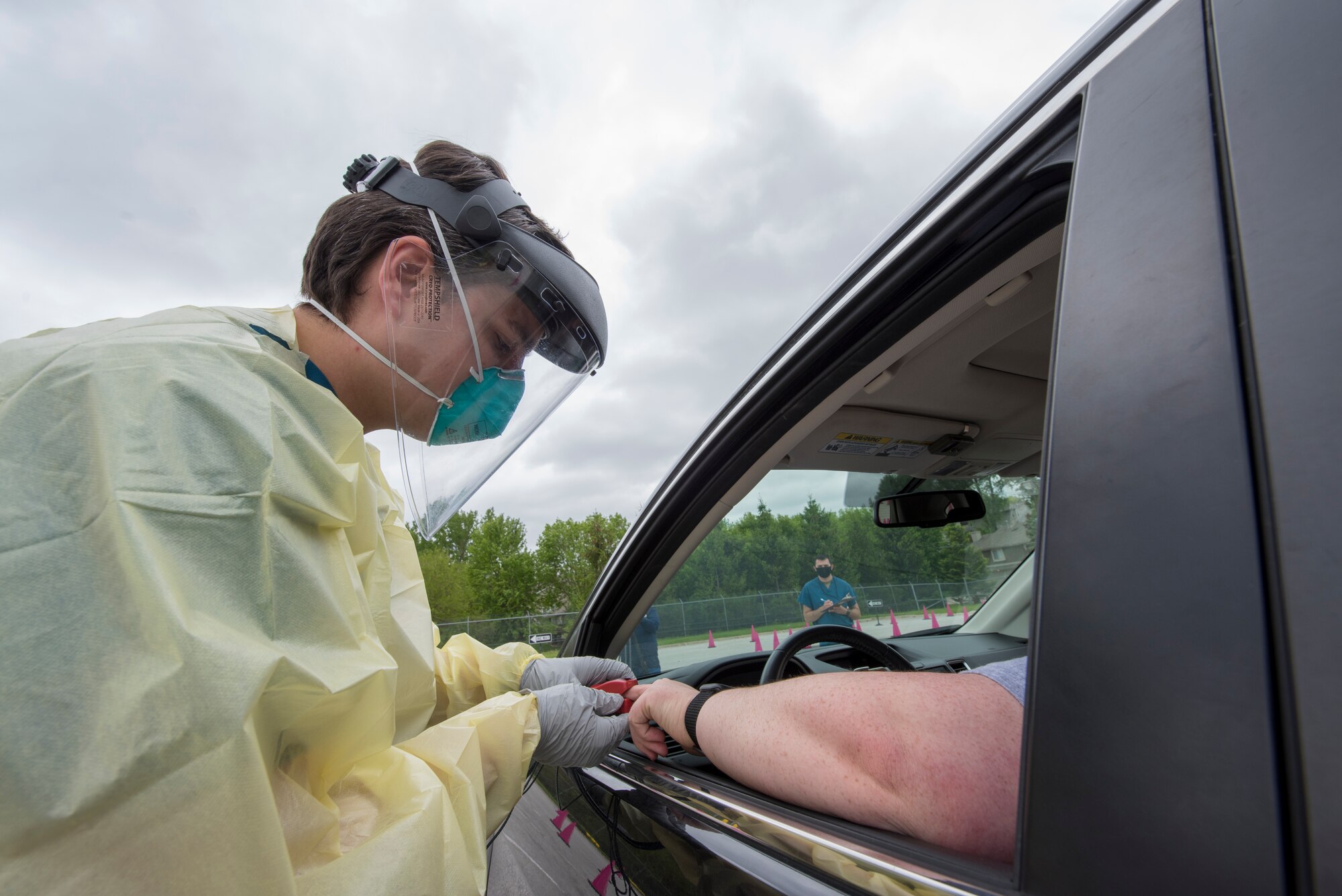 service member in PPE puts clip on patients finger through car window.