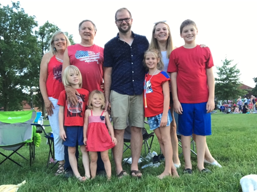 Two grandparents, a husband and wife and their four kids smile wearing red, white and blue at a 4th of July Ceremony.