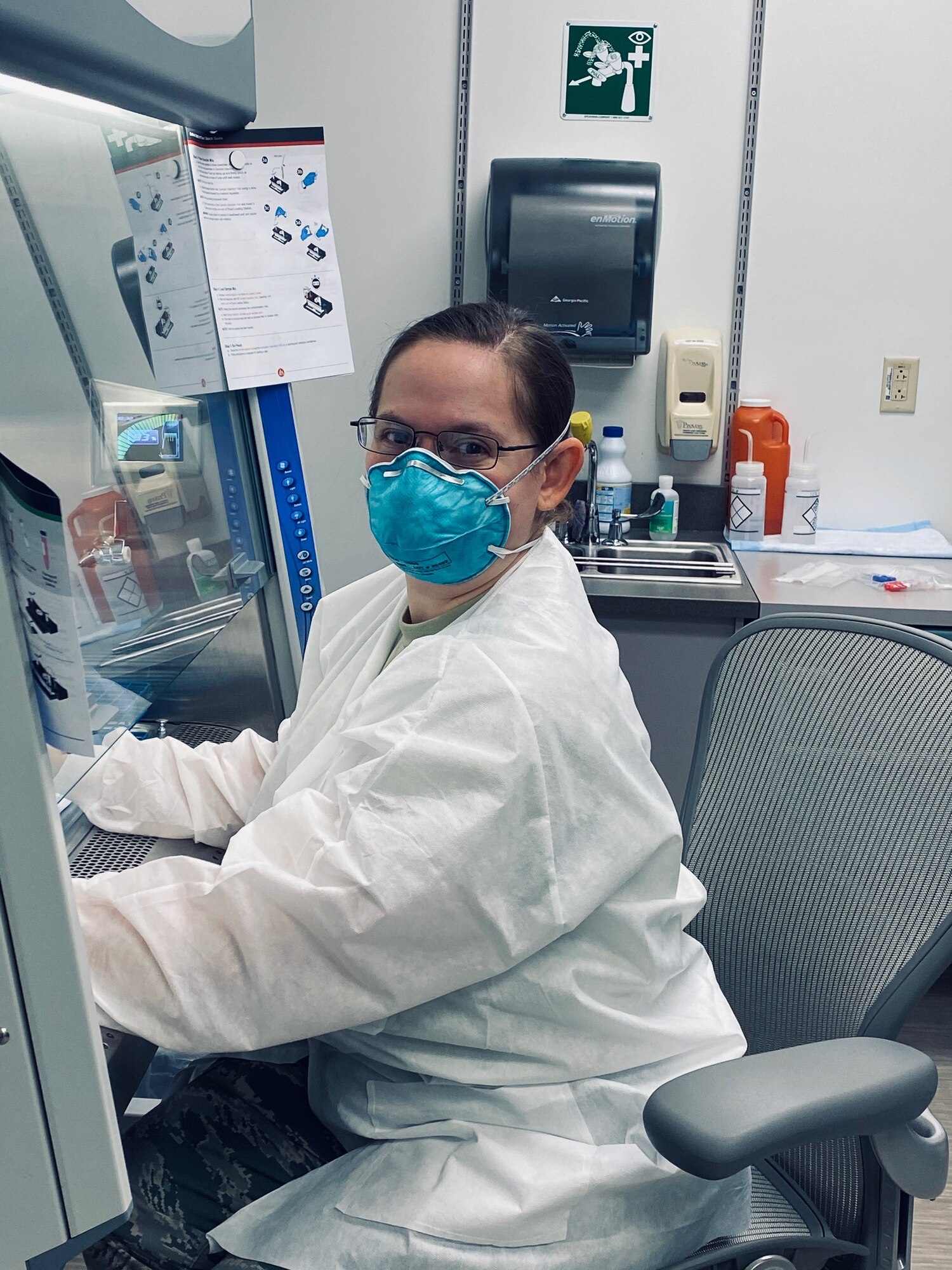 Photo shows woman in hospital gown and mask working on a large laboratory machine.