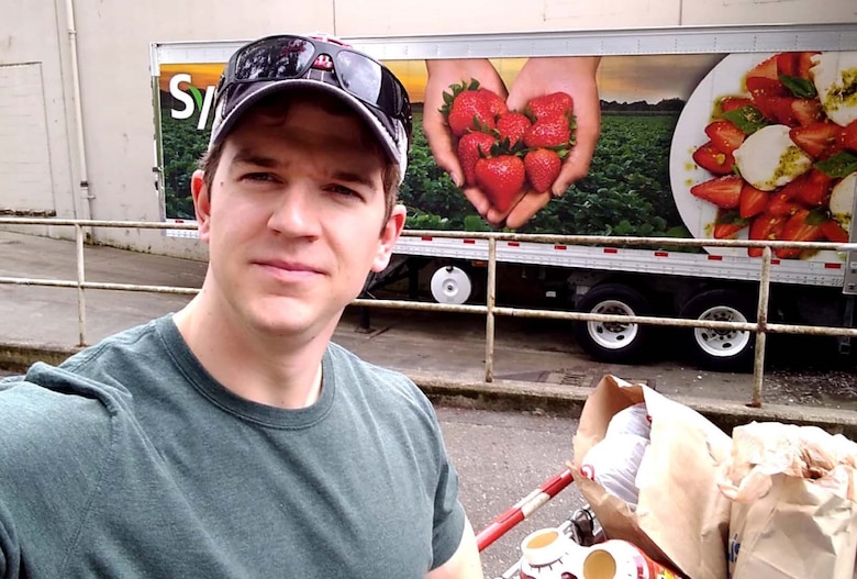 A Caucasian male stands in front of a food delivery truck along with paper bags filled with supplies.