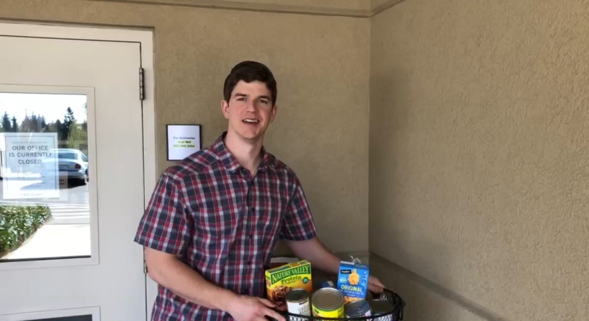 A Caucasian male stands in front of a beige building with a bag filled with food and supplies for COVID-19 relief.