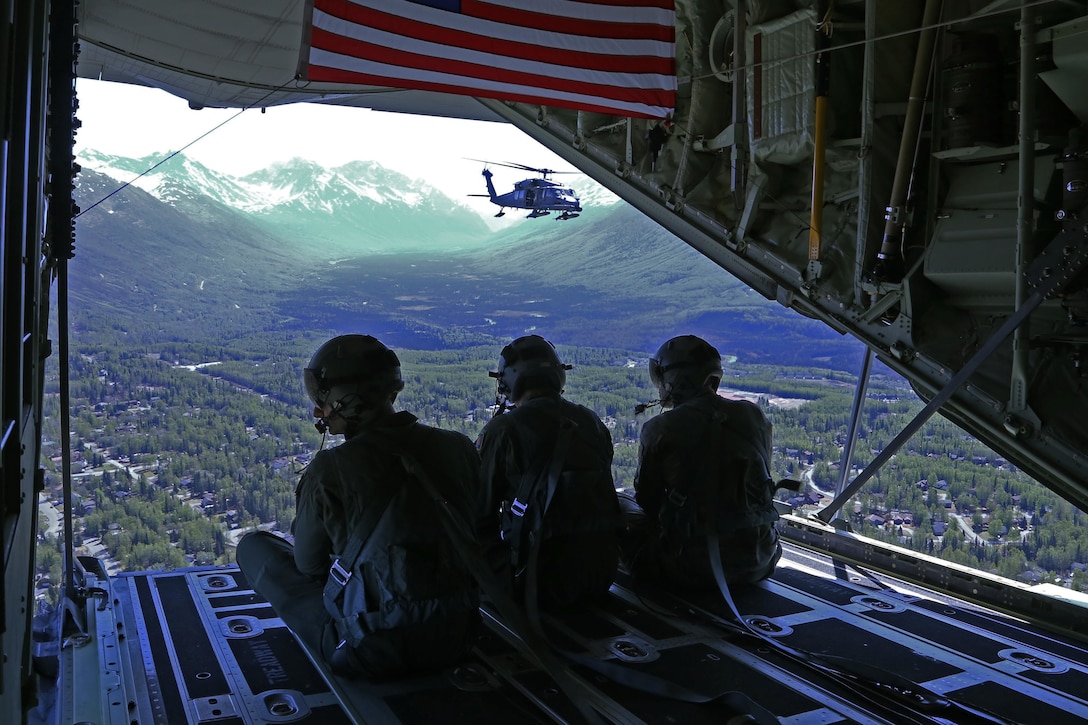Three service members sit in an open part of a large military aircraft in flight looking at a helicopter.
