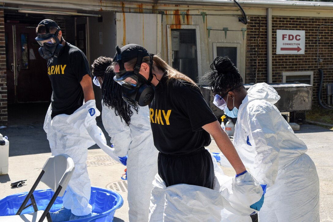 Outside, two women dressed in protective clothing help a man and a woman remove their protective clothing.