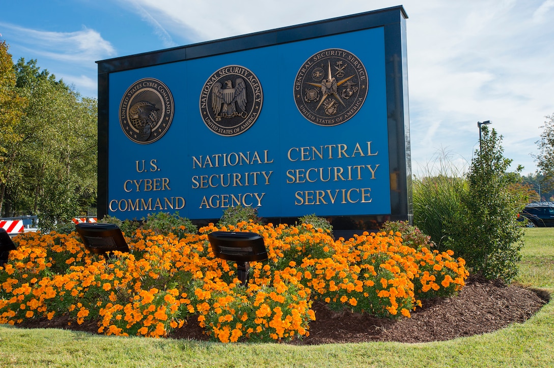 Three seals are affixed to an outdoor sign that identifies each seal:  “U.S. Cyber Command,” “National Security Agency,” and “Central Security Service.” Brightly colored flowers bloom around the sign..
