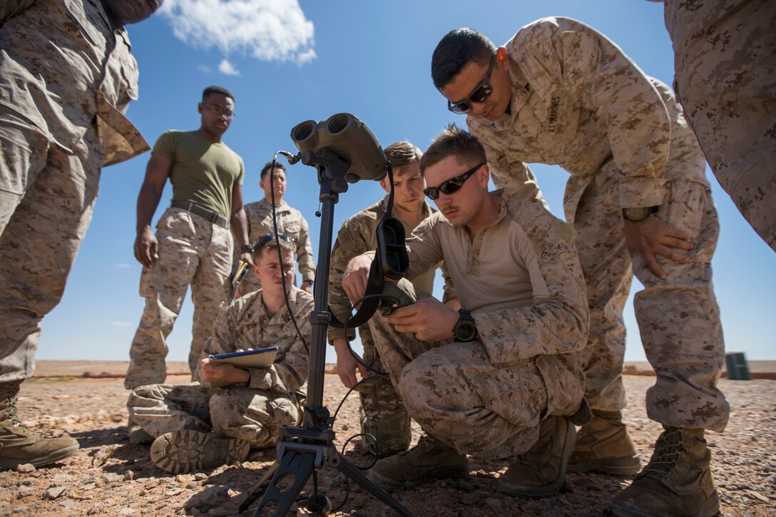 Marines gather around an airman holding GPS equipment and kneeling outside on desert-like terrain.