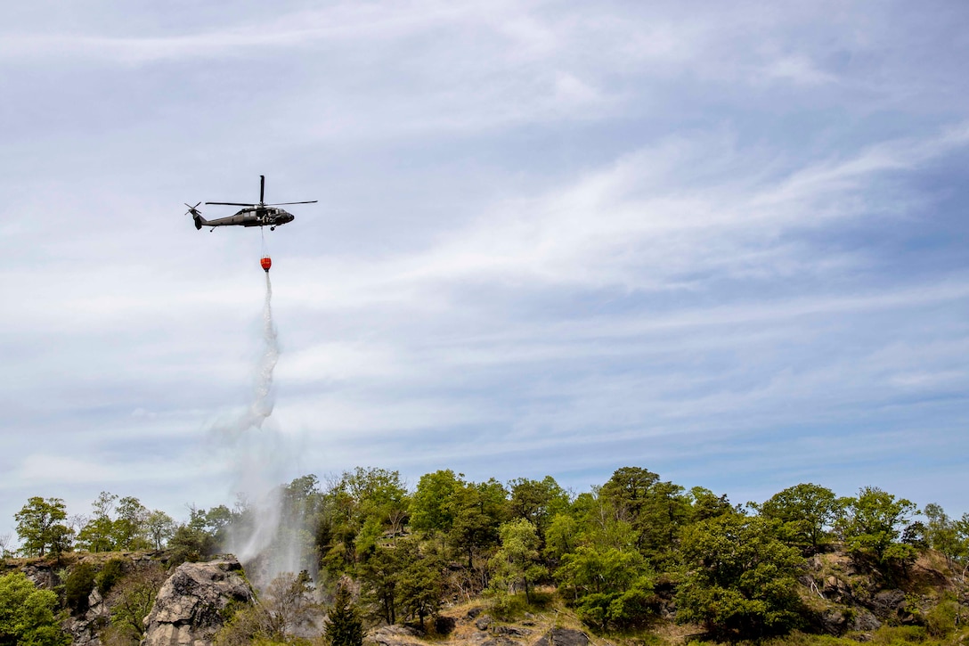 An Army helicopter drops water from a bucket over a tree line.