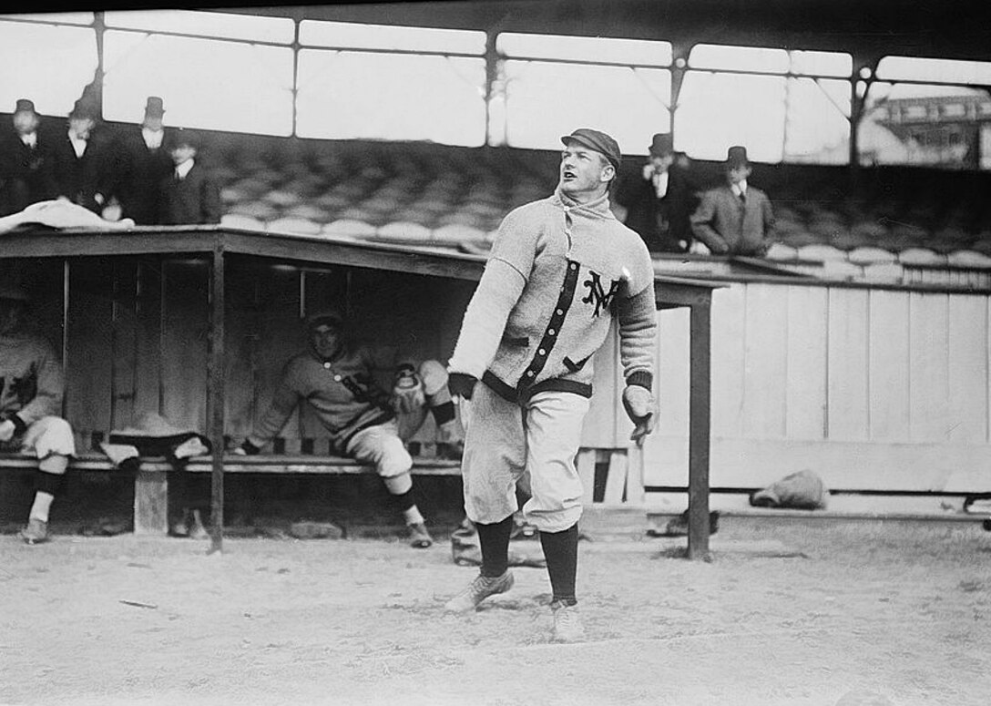 A baseball pitcher wearing a New York Giants sweater warms up in front of a dugout.
