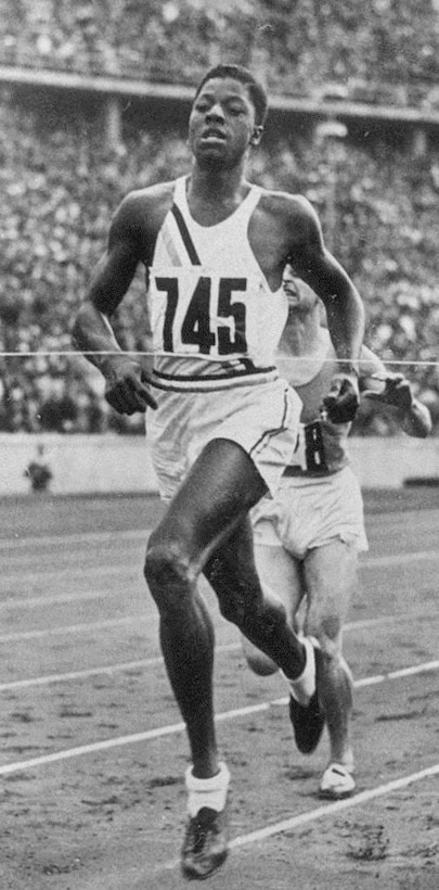 Running athlete just seconds before breaking the tape at the finish line. A packed grandstand is in the background.