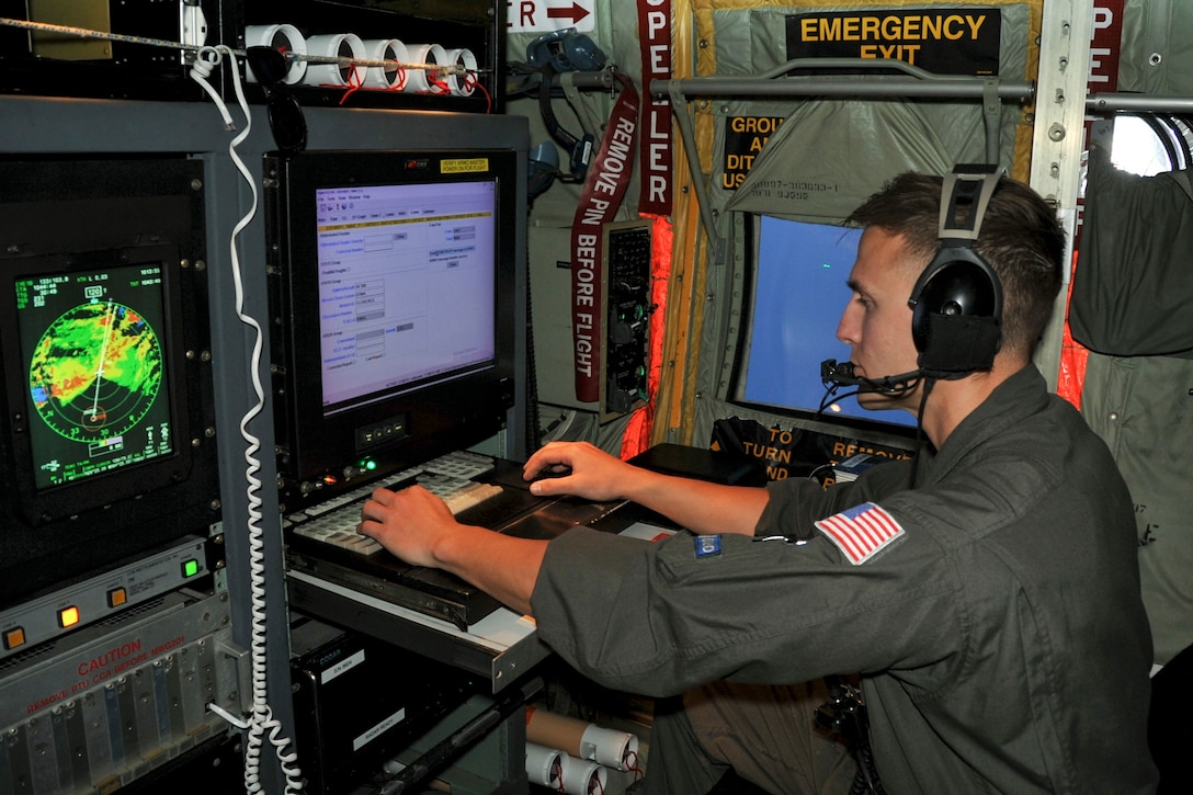 An airman sits in front of screens displaying data at a workstation inside an aircraft.