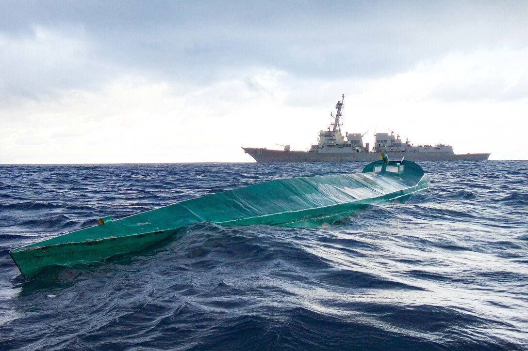 In the foreground, a drug-running vessel at sea with a U.S. guided-missile destroyer in the background.