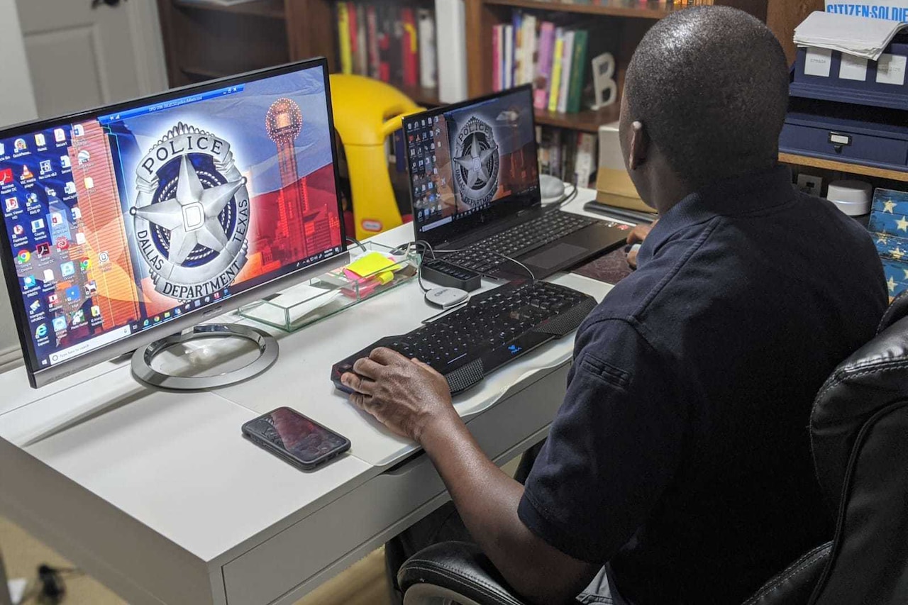 A man at a desk observes two computer screens while using a computer.