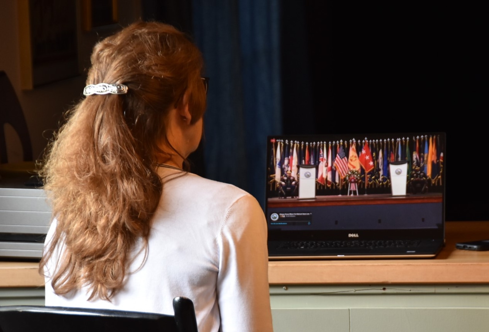 A woman is seated watching a streaming video of a keynote address from a laptop