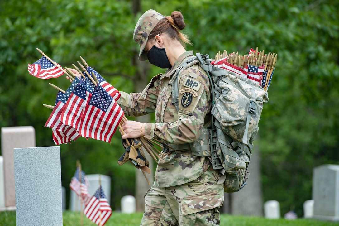 A soldier wearing a face mask walks through a cemetery carrying small American flags.