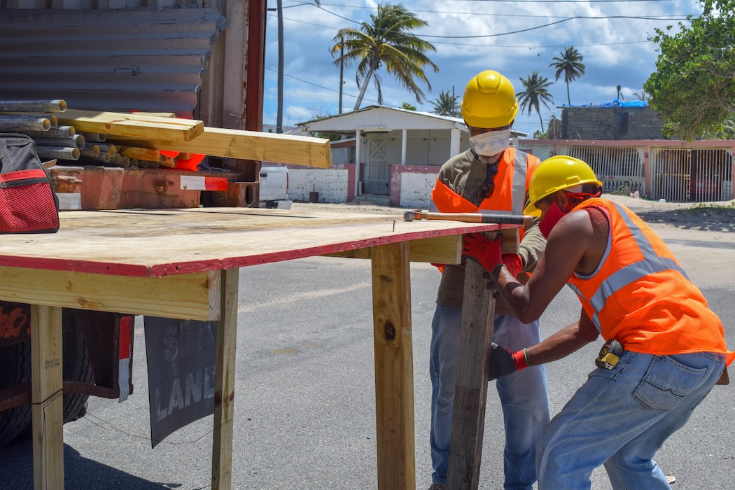 Two men wearing yellow helmets and orange safety vest put together a wooden structure