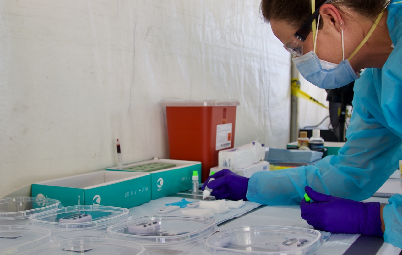 A nurse wearing protective gear leans over a counter working with lab materials.