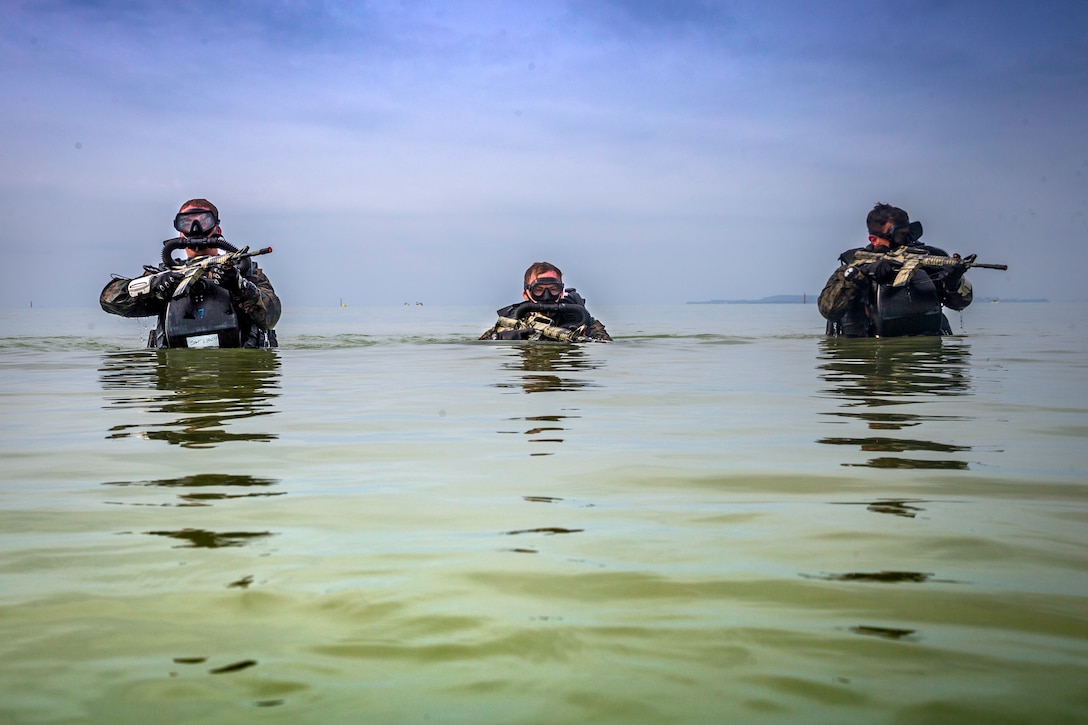 Three Marines wade through water carrying guns.