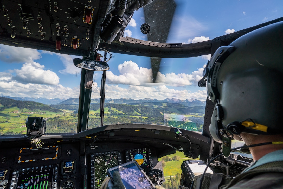A solider holds a digital device and looks out from a cockpit over mountains.