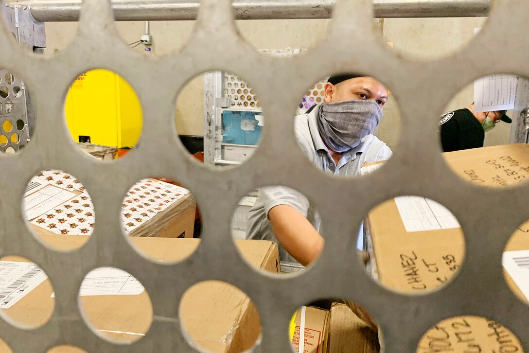 A civilian mail clerk wearing a mask loads packages into a postal bin at a Navy mail facility.