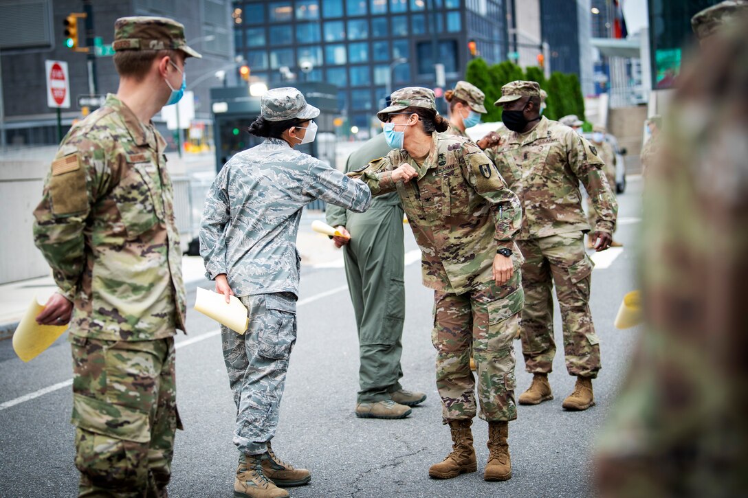 Service members wearing face masks and observing social distancing greet each other by touching elbows.