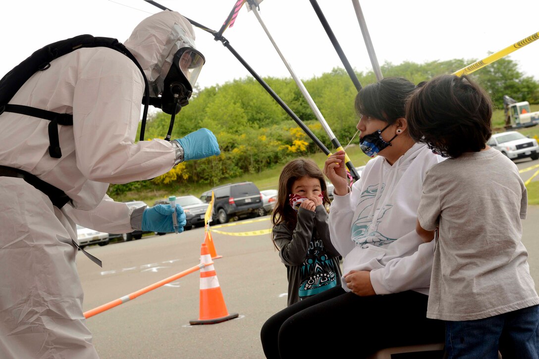 An airman wearing personal protective equipment stands in front of a woman and two children as the woman holds a swab toward her nose.