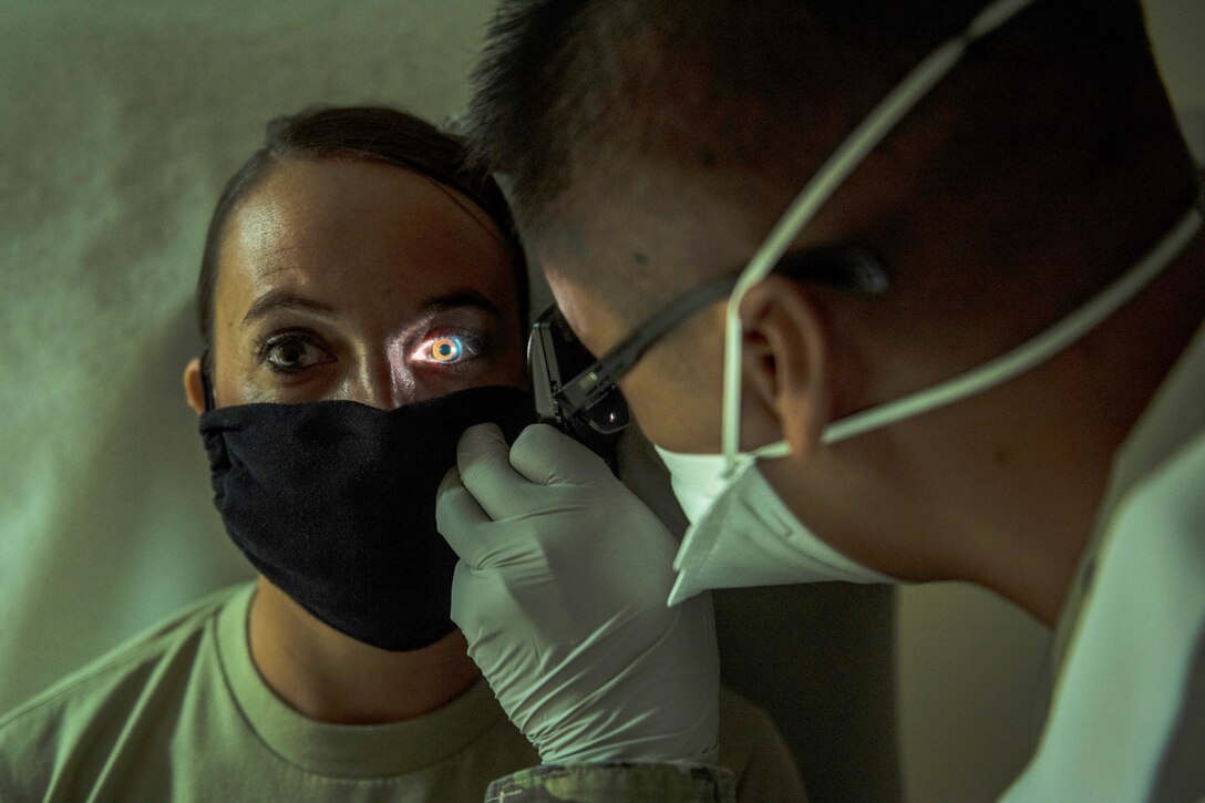 An Air Force physician in a face mask uses an instrument with a light to examine the eyes of a patient, who is also wearing a mask.