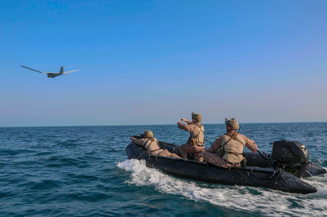 Three Marines ride in a combat rubber raiding craft; an unmanned aircraft system seen in the distance.