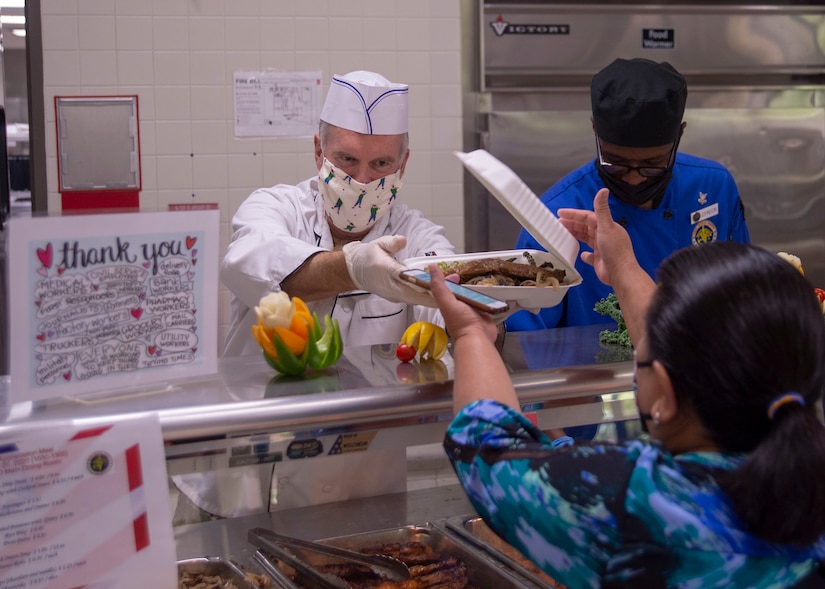 A sailor wearing a face mask hands off a container of food over a counter.