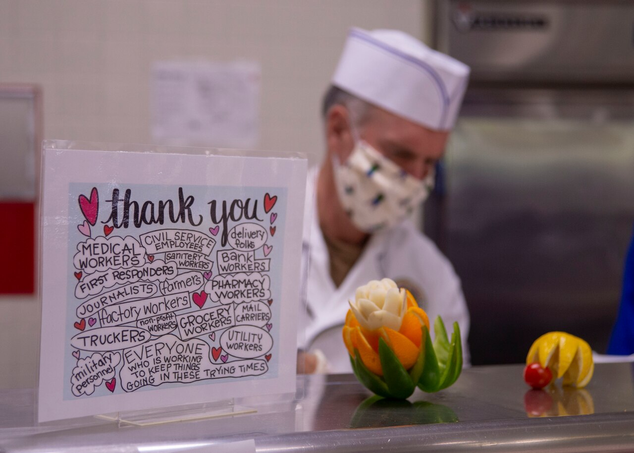 A sailor wearing a face mask stands behind a Thanks You sign sitting on a counter.