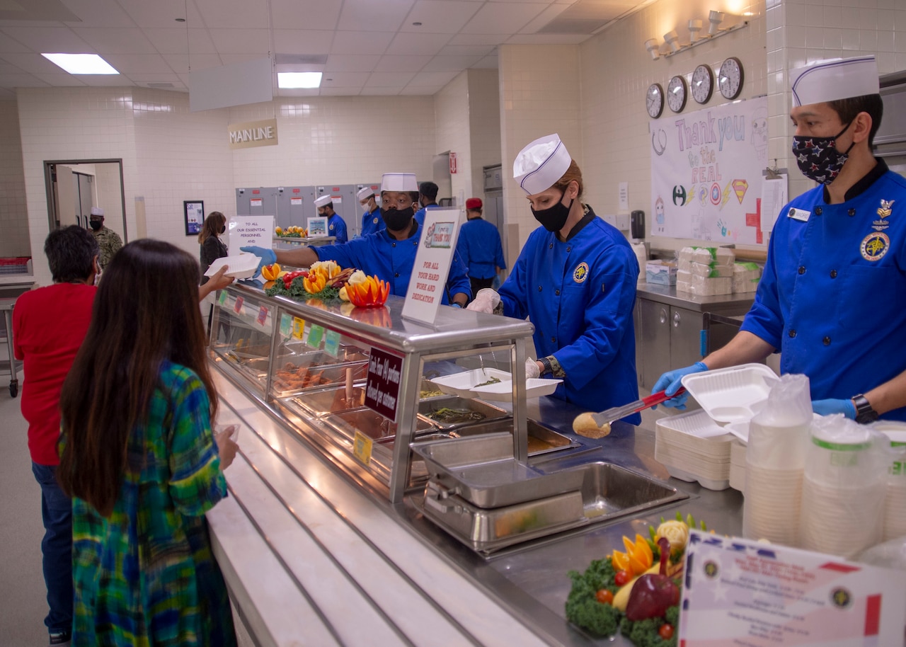 Sailors wearing gloves and face masks serve food.