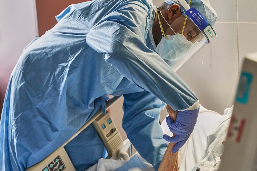 A sailor wearing protective gear holds the wrist of a patient in a bed.