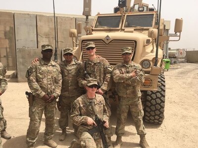 six men in green camouflage uniforms and caps with 9mm pistols in holsters on sides and one holds an M4 rifle stand in front of a military vehicle.