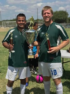 Hispanic male and white male in green jerseys and white shorts hold one large trophy and two little trophies.