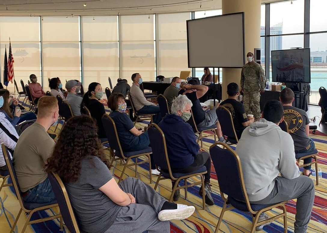 A soldier stands at the front of a room full of people seated in chairs.