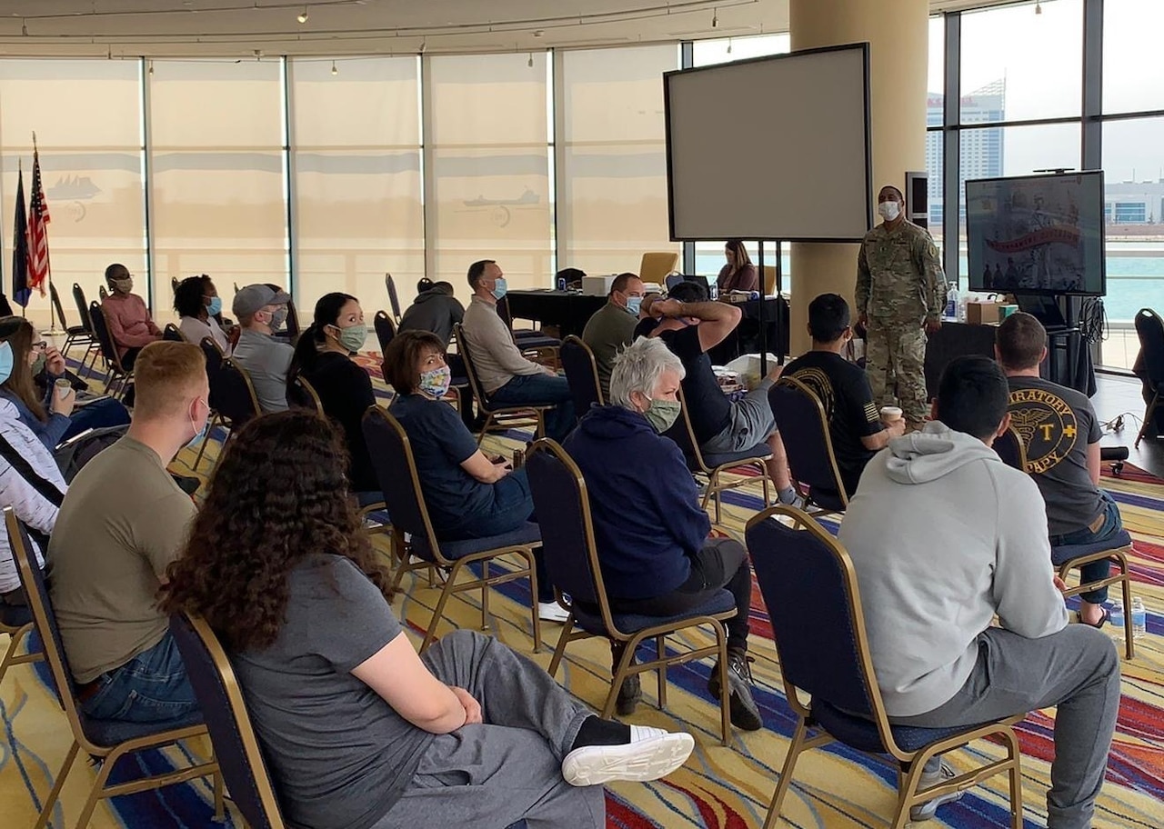 A soldier stands at the front of a room full of people seated in chairs.