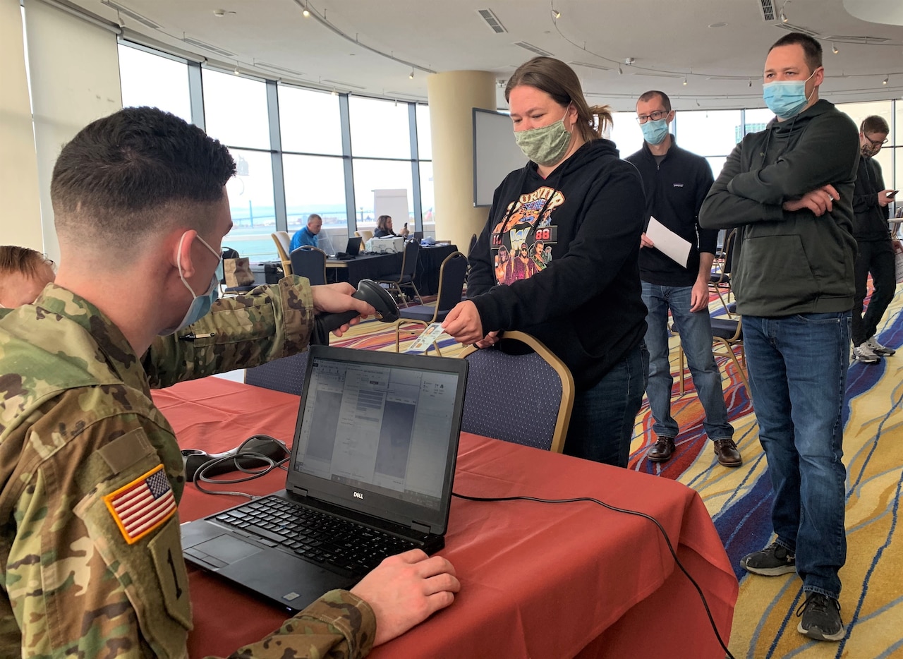 A soldier sits at a laptop computer taking identification cards from a line of people.