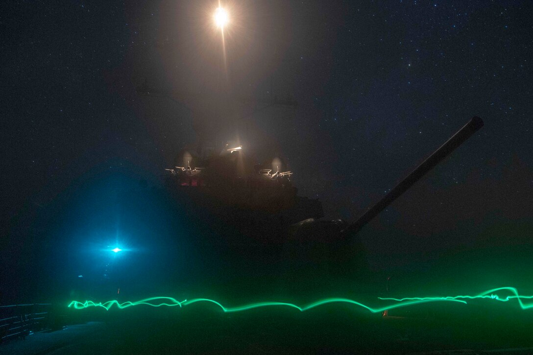 A Naval ship transits a body of water illuminated by a green light.