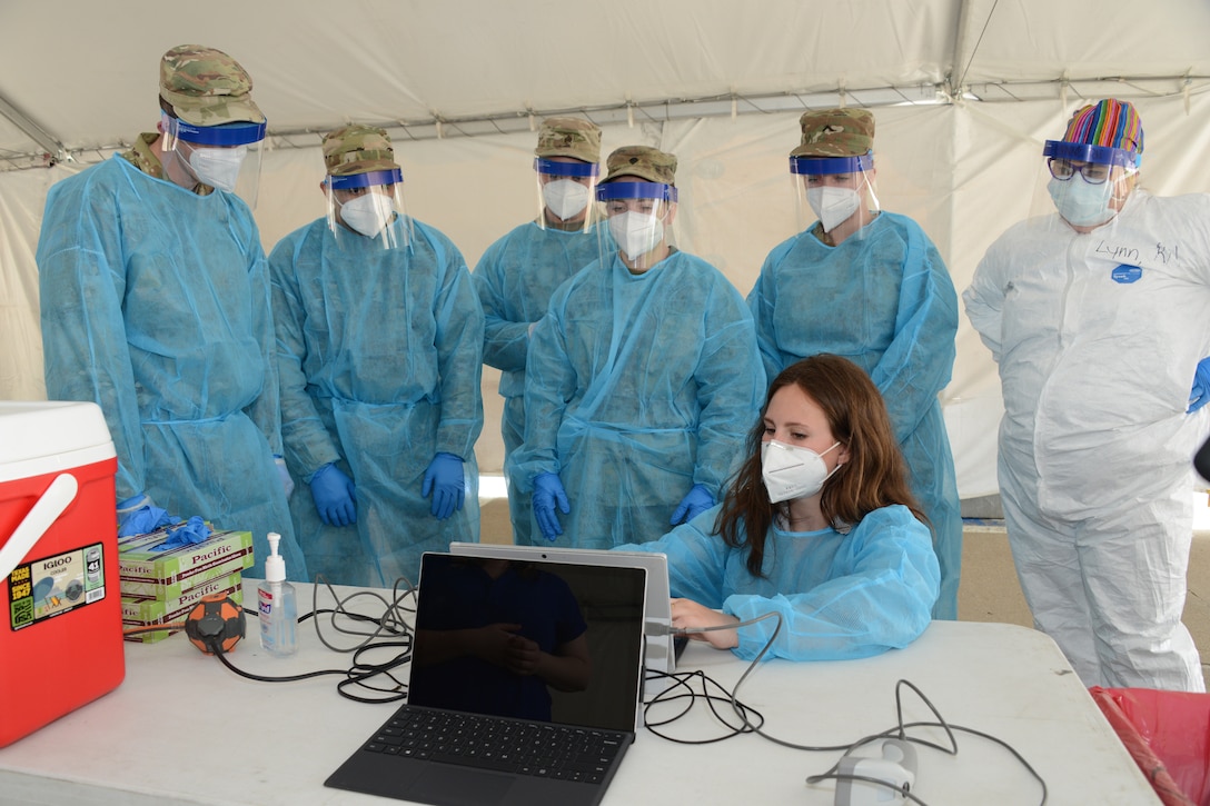 A group of National Guardsmen in medical uniforms stand behind a woman on a computer.